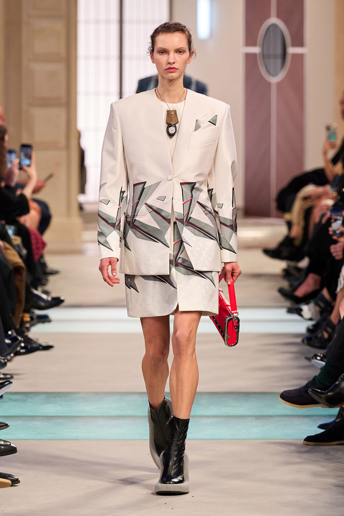 Female model on a fashion runway wearing a white blazer and matching shorts with a geometric gray and red print, paired with black ankle boots and a small red handbag. Audience members are seen watching and taking photos in the background.