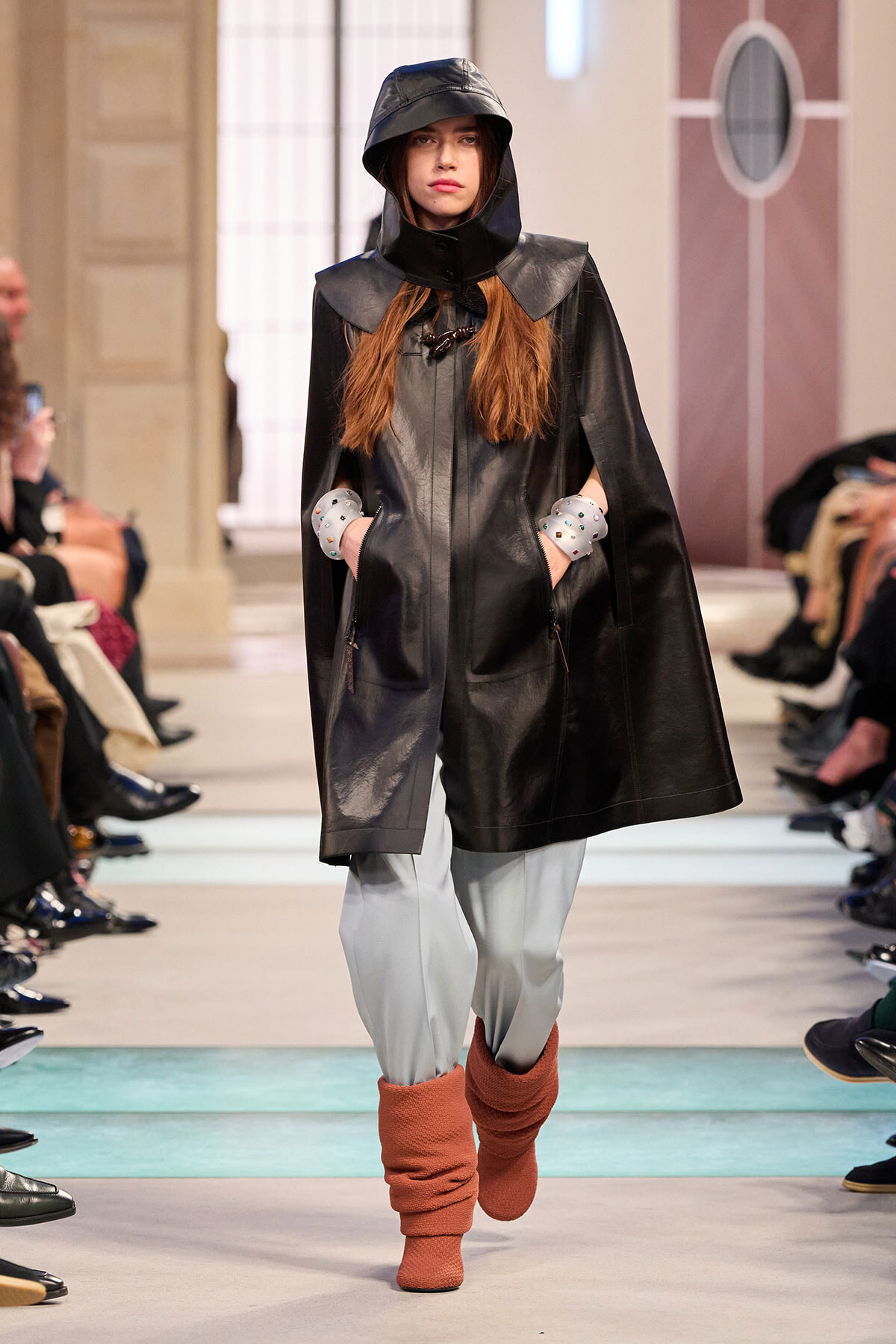 Model walks the runway in a black hooded leather cape with exposed shoulders and hands in pockets, paired with light gray pants and orange knit leg warmers.