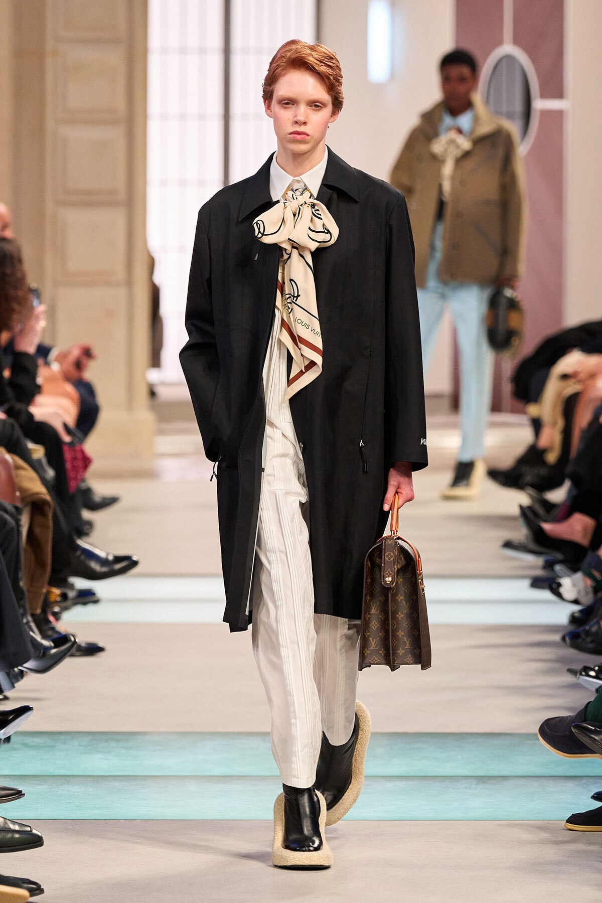 Model walks the runway in a black coat with a large cream scarf, white striped pants, and carries a Louis Vuitton handbag.