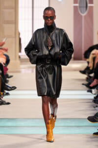 Model on a runway wearing an oversized black leather jacket with a matching knee-length skirt and a belt, tan ankle boots, and dark sunglasses, accessorized with a pendant bag charm.