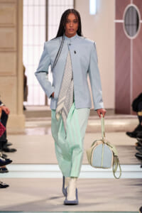 Model walking a runway in a pale blue blazer and mint trousers, carrying a pastel handbag and scarf-wrapped outfit accessory.