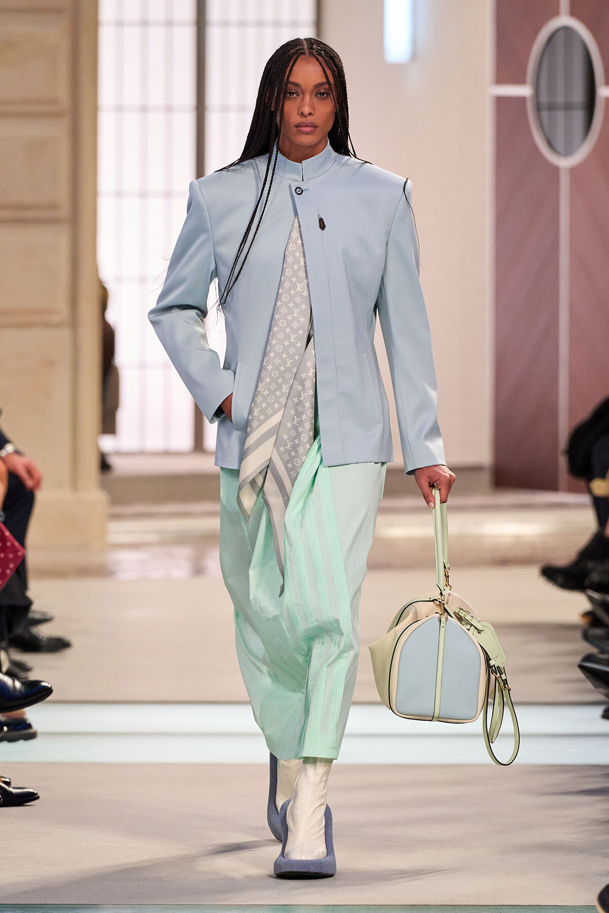 Model walking a runway in a pale blue blazer and mint trousers, carrying a pastel handbag and scarf-wrapped outfit accessory.