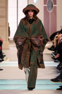 Runway model in an oversized green-brown cape coat with plaid lining and two Louis Vuitton belt bags, wearing a wide-brim hat.