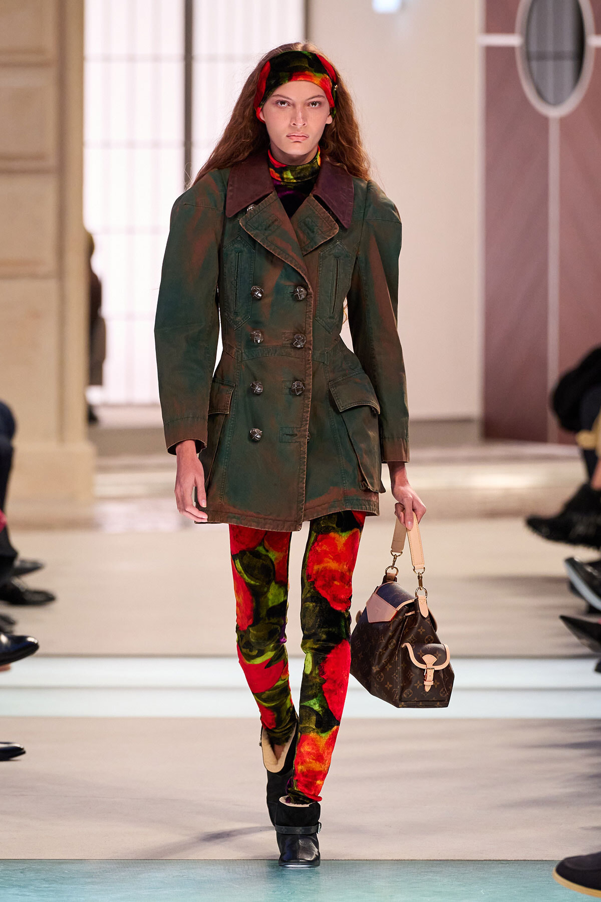 Model on a runway wearing a green double-breasted coat, colorful tie-dye leggings, a red headband, and a brown designer handbag.