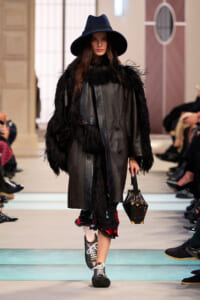 Model walks a runway in a black fur-trimmed coat and wide-brim hat, carrying a structured handbag.