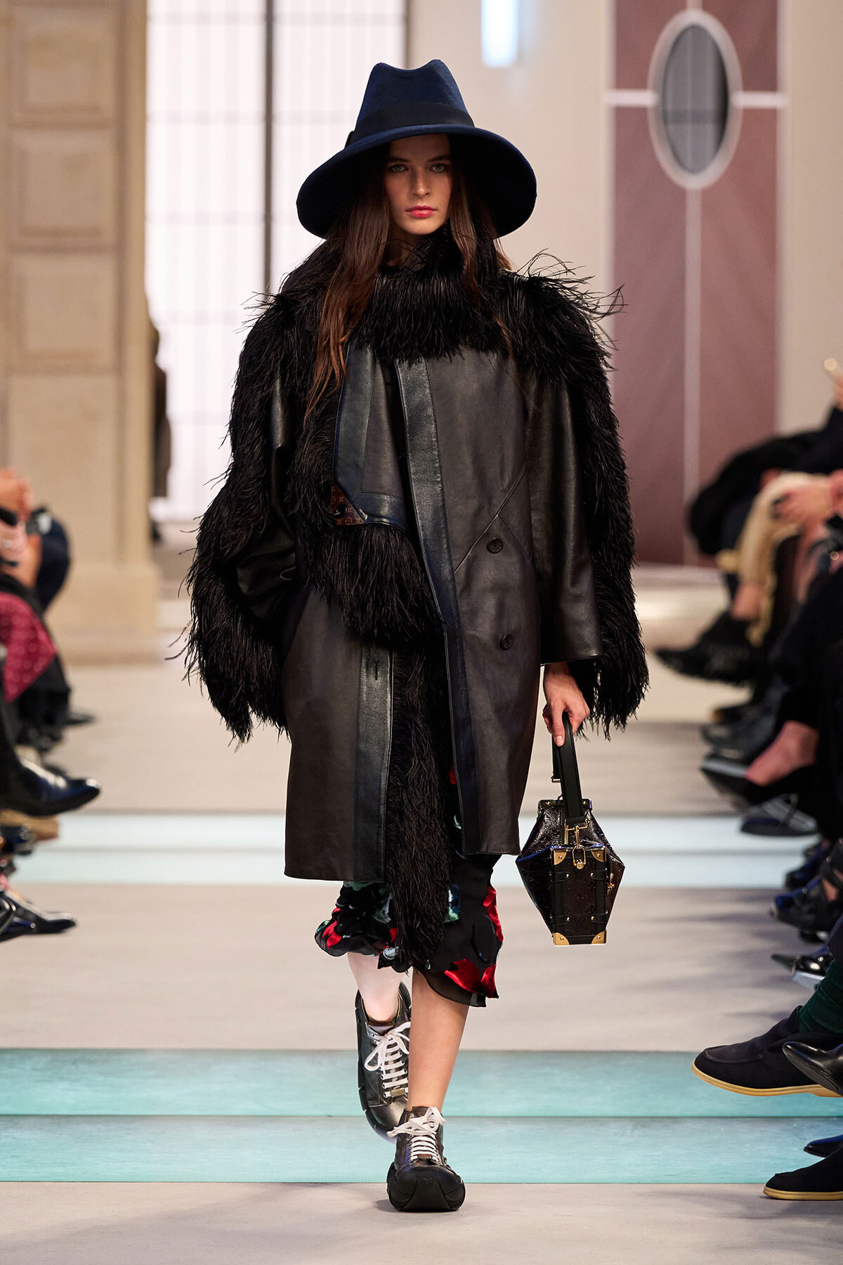 Model walks a runway in a black fur-trimmed coat and wide-brim hat, carrying a structured handbag.
