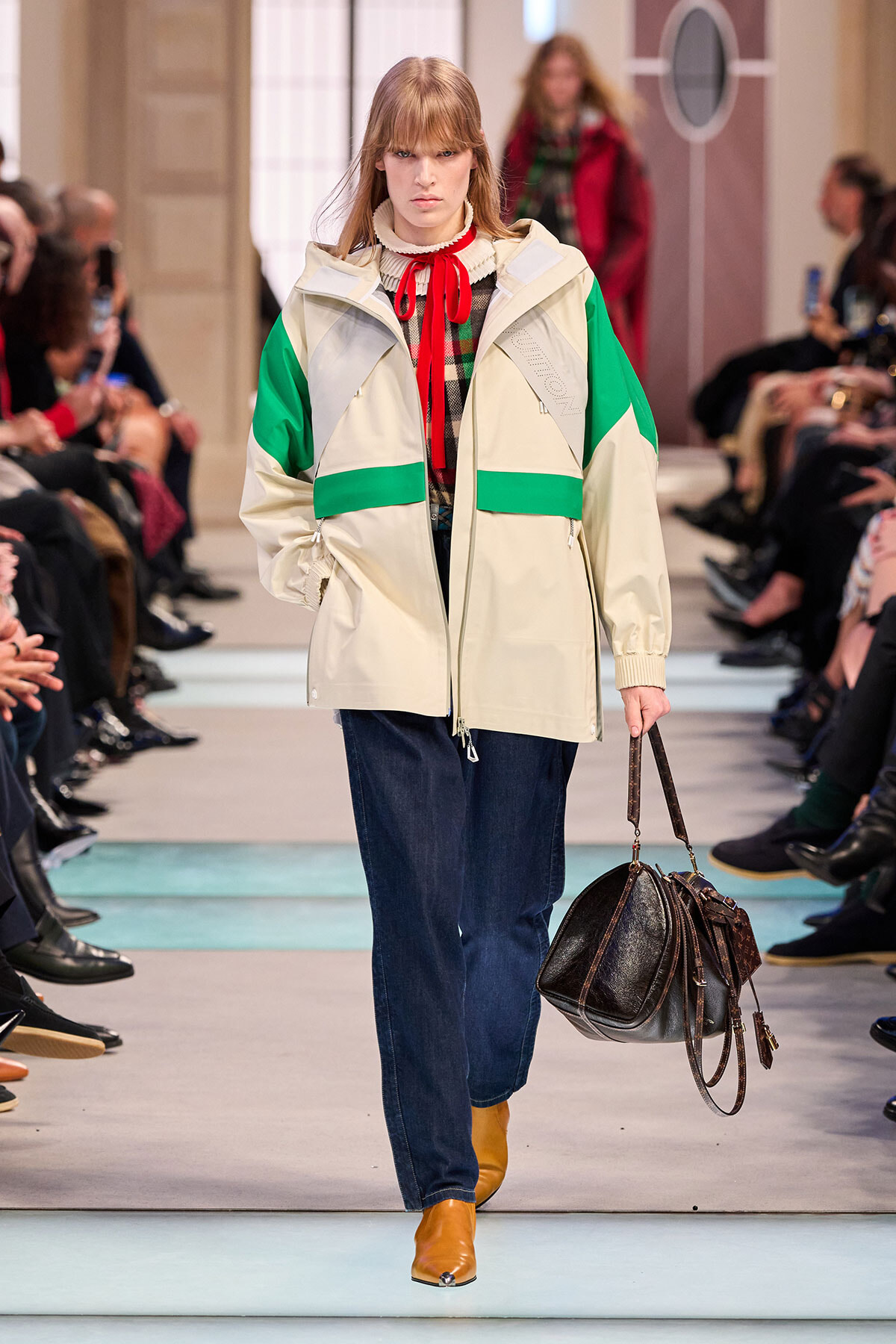 Model walks the runway in a beige oversized jacket with green color blocks, a plaid shirt featuring a red tie, loose blue jeans, and tan ankle boots, carrying a brown leather duffel bag.