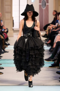Model walking a runway in a black tiered ruffle skirt, black tank top, wide belt, and oversized black hat, carrying a handbag.