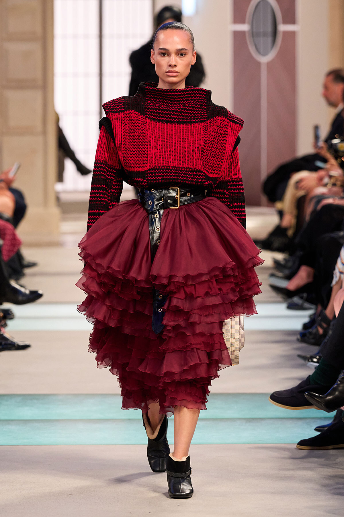Model walking a runway in a red-and-black chunky knit top and a voluminous burgundy tiered skirt with a wide black belt.