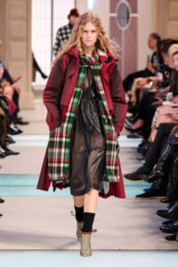 Female model walking a runway in a burgundy coat, black dress, and a green plaid scarf during a fashion show.
