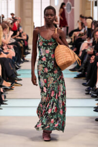 Model walks a floral green maxi dress down a fashion runway, carrying a tan woven handbag and wearing pink shoes as spectators look on.