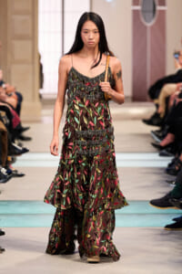 Model walking a fashion runway in a sheer, layered dress with colorful leaf patterns and a tan shoulder bag