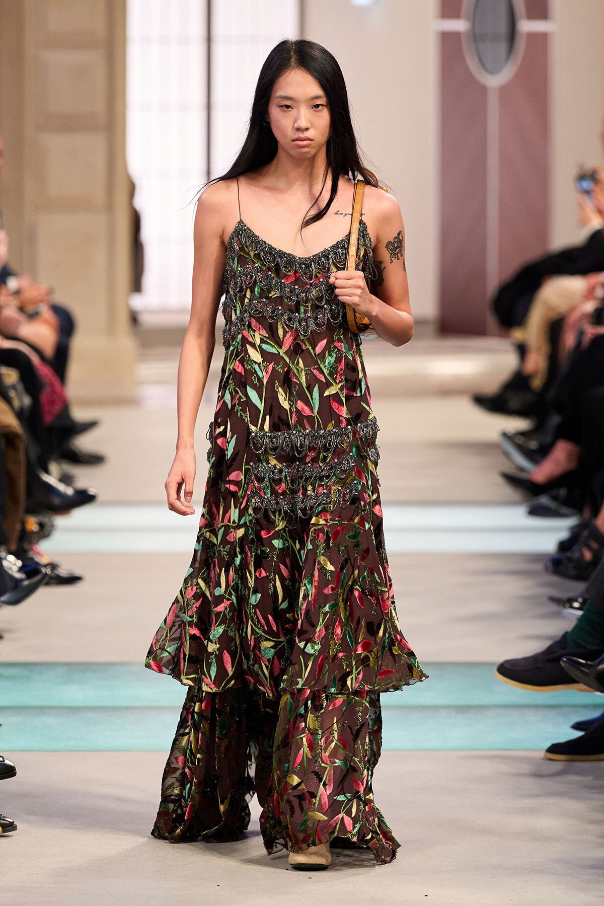 Model walking a fashion runway in a sheer, layered dress with colorful leaf patterns and a tan shoulder bag
