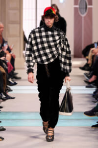 Model walks the runway in a bold black-and-white checkered cape draped over one shoulder, with a red cap, black velvet pants, and a two-tone black-and-white handbag.