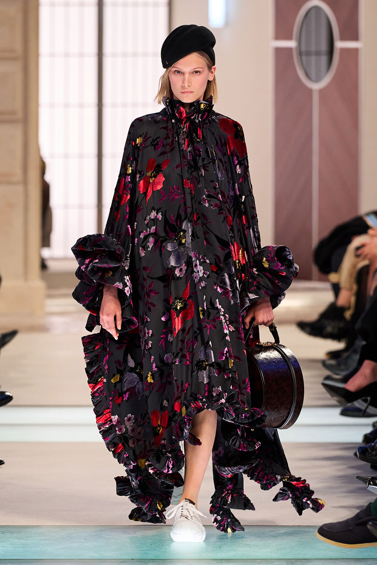 Model walks runway in a voluminous black floral gown with ruffled sleeves, beret, and white sneakers, carrying a round dark instrument case.