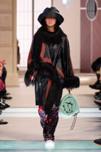 Model walking the runway in a black leather coat with fur trim and red panels, paired with patterned pants, a wide-brim hat, and a mint green bag.