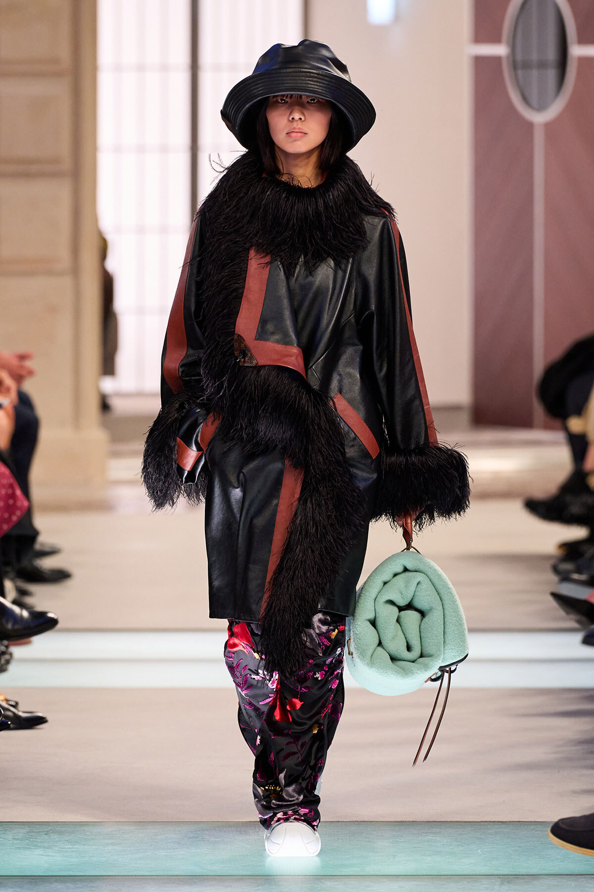 Model walking the runway in a black leather coat with fur trim and red panels, paired with patterned pants, a wide-brim hat, and a mint green bag.