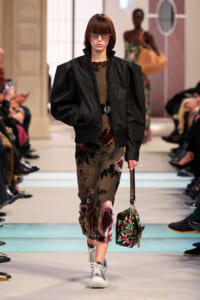 Model on a runway wearing a black leather bomber jacket over a floral midi dress, with sunglasses and a small floral handbag.