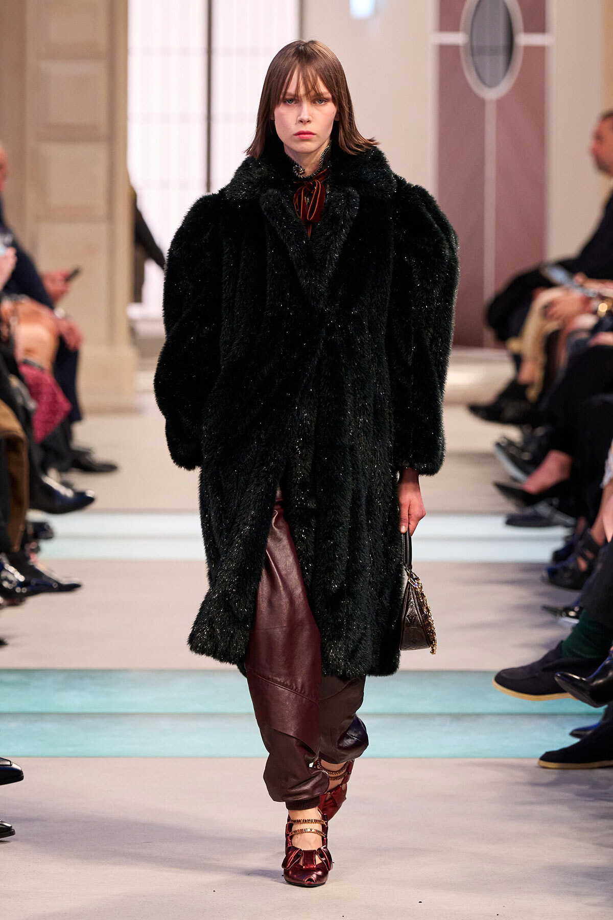 Model walks a fashion runway wearing a long black faux fur coat, burgundy leather pants, and red strappy shoes as the audience watches.