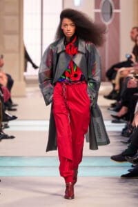 Model walking a runway in a bold red-and-gray outfit: red pants, colorful top, oversized gray coat, red scarf, brown ankle boots, with audience watching.