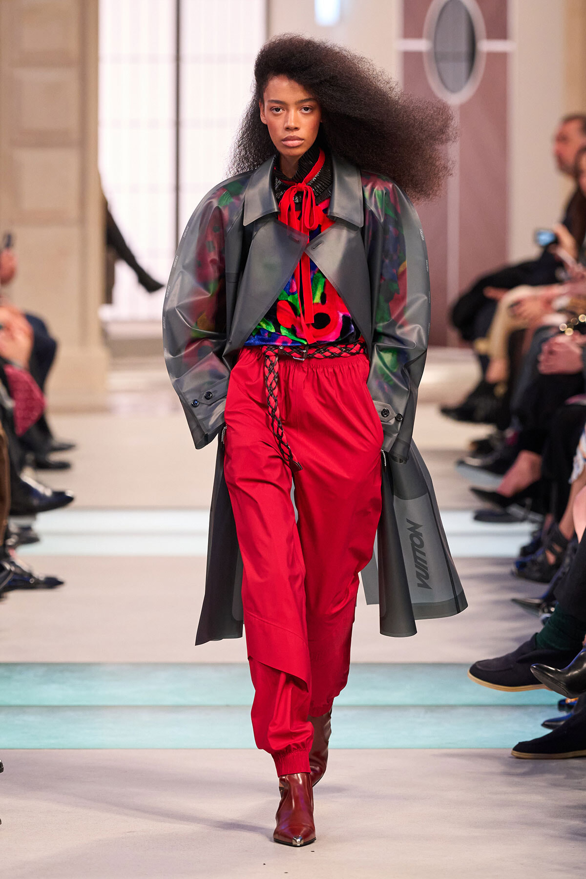 Model walking a runway in a bold red-and-gray outfit: red pants, colorful top, oversized gray coat, red scarf, brown ankle boots, with audience watching.
