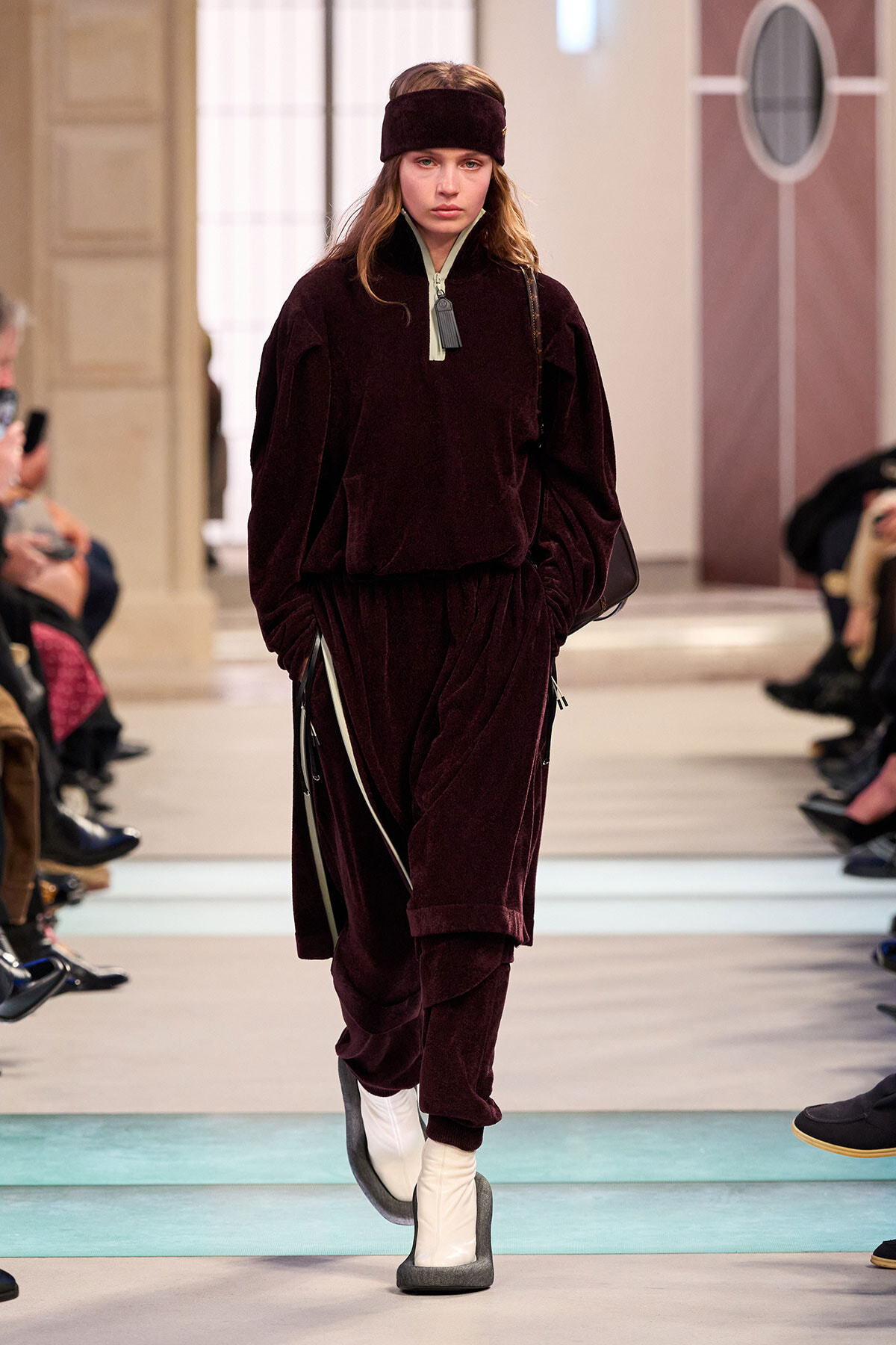 Model on a runway wearing a burgundy velour tracksuit with matching headband and white boots in a fashion show setting.