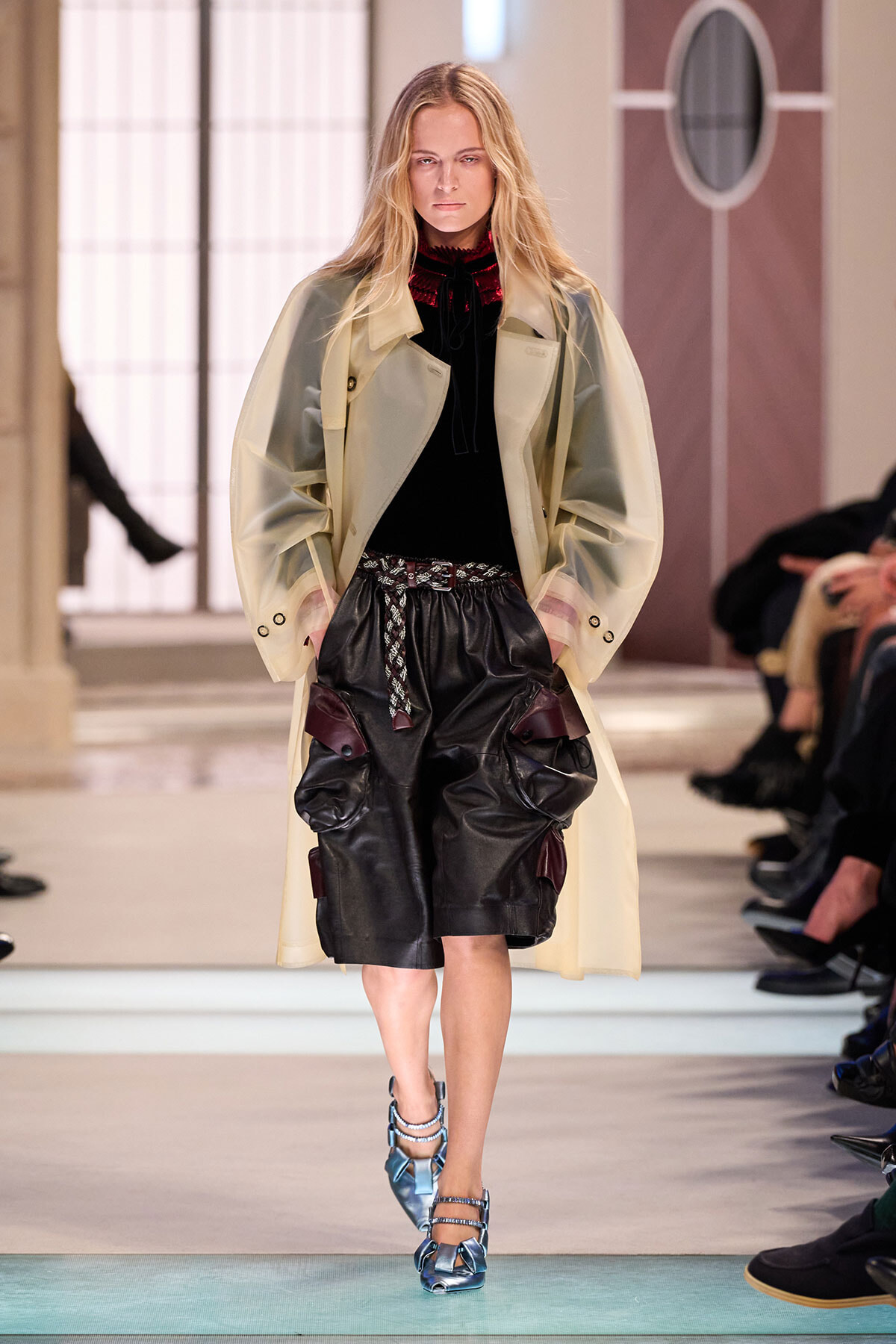 Model walks the runway wearing a long pale beige trench over a black top and oversized black leather shorts with a rope belt, metallic blue strappy heels, in a fashion show setting.