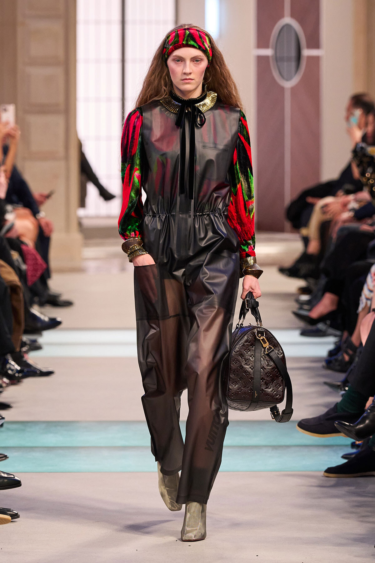 Model walks a runway in a shiny black jumpsuit with red-green velvet sleeves, gold cuffs, and a matching headband, carrying a brown quilted bag.