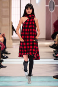 Model walking the runway in a red and black plaid halter dress with a scarf, wearing knee-high socks and silver ankle boots.