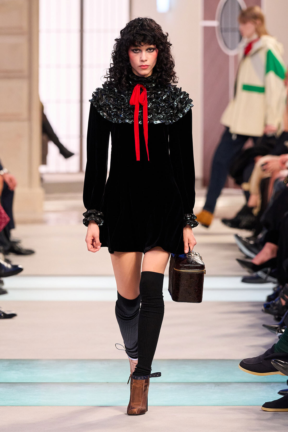 Model walking a fashion runway in a black velvet dress with a ruffled collar and red ribbon, carrying a small brown hatbox-style bag.