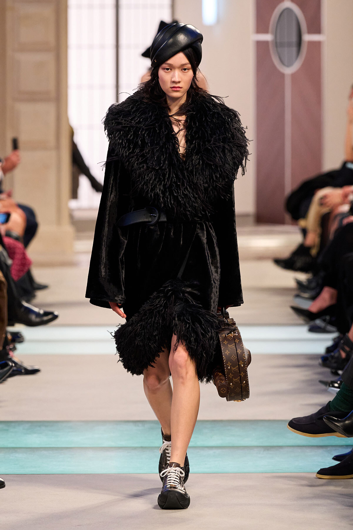 Model on a fashion runway wearing a black fur coat and a black turban-like hat, carrying a brown Louis Vuitton suitcase.