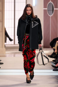 Model walking a runway in a black oversized jacket with a Vuitton logo, paired with red-green velvet pants and black strappy shoes, carrying a light beige bag.