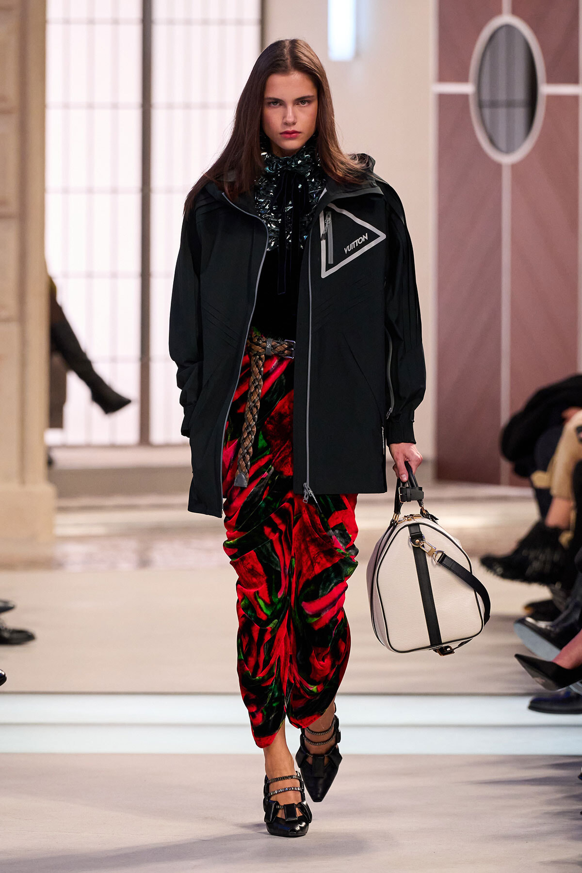 Model walking a runway in a black oversized jacket with a Vuitton logo, paired with red-green velvet pants and black strappy shoes, carrying a light beige bag.