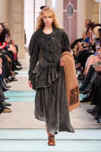 Model walking the runway in a dark denim-gray layered dress, holding a camel coat with a LV-patterned pocket.