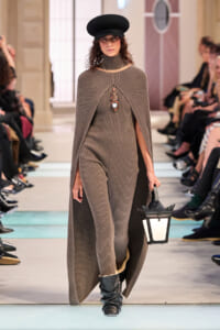 Model walking a fashion runway in a taupe ribbed knit cape dress, black hat, and knee-high boots, carrying a lantern.