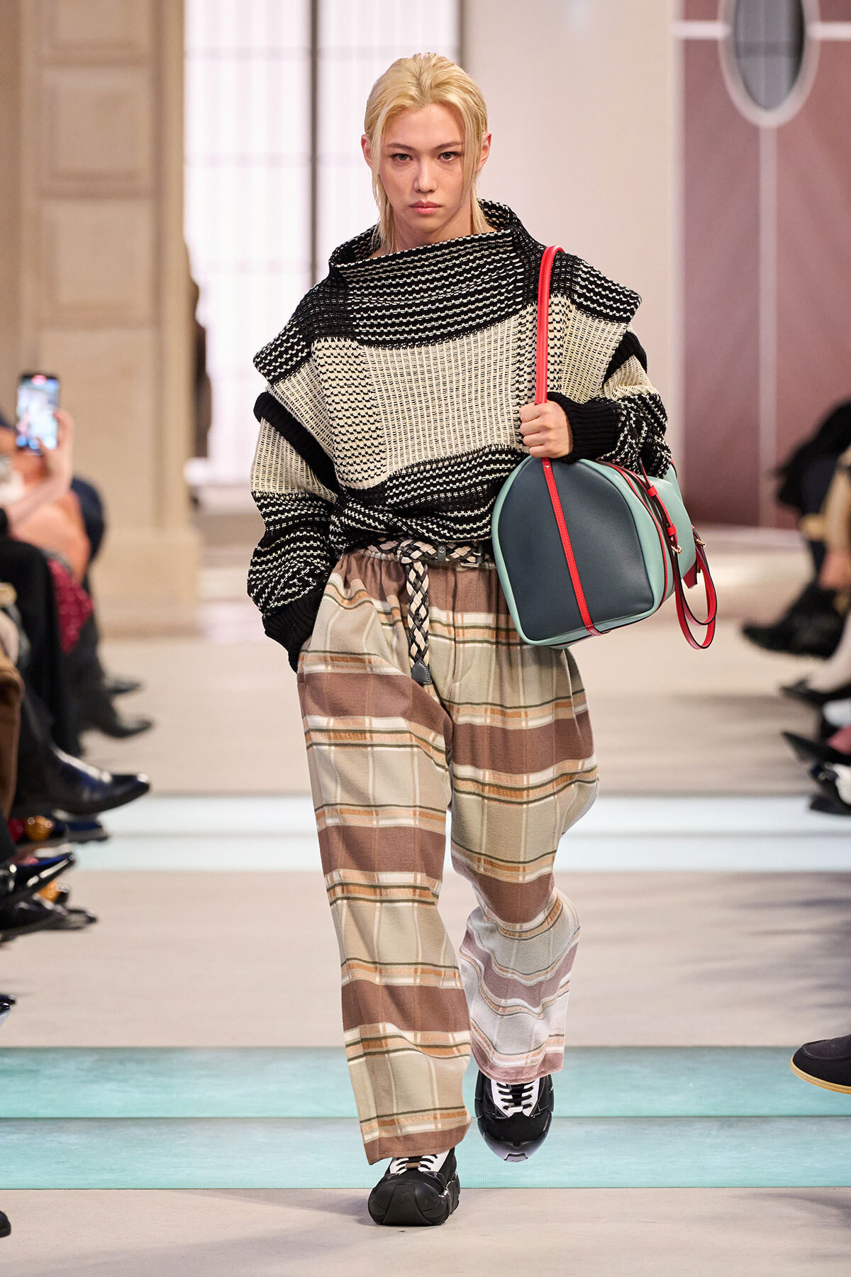 Model on a runway wearing an oversized black-and-white knit sweater, beige plaid wide-leg pants, and a teal bag with a red strap caped over the shoulder.