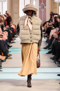Model on a runway wearing a cream fuzzy puffer vest over a dark sweater, a beige maxi skirt, brown ankle boots, and a wide-brim hat with a scarf tied at the neck.
