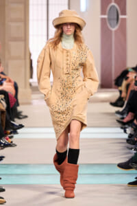 Model on a runway wearing a beige, embellished dress with turquoise beads, a wide-brim hat, and rust-colored slouch boots.