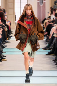 Female model walking a runway in an oversized brown coat over a red striped turtleneck, with a pale green skirt and chunky knit gloves, audience along the sides.