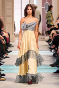 Model on a runway wearing a cream satin gown with a gray draped bodice and layered gray fringe at the hem, audience seated on sides.