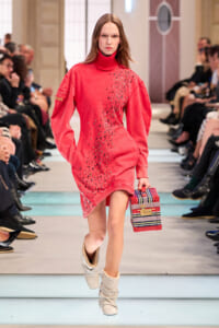 Model walking a fashion runway in a red turtleneck sweater dress with scattered black embellishments, carrying a striped box handbag and wearing light beige boots.