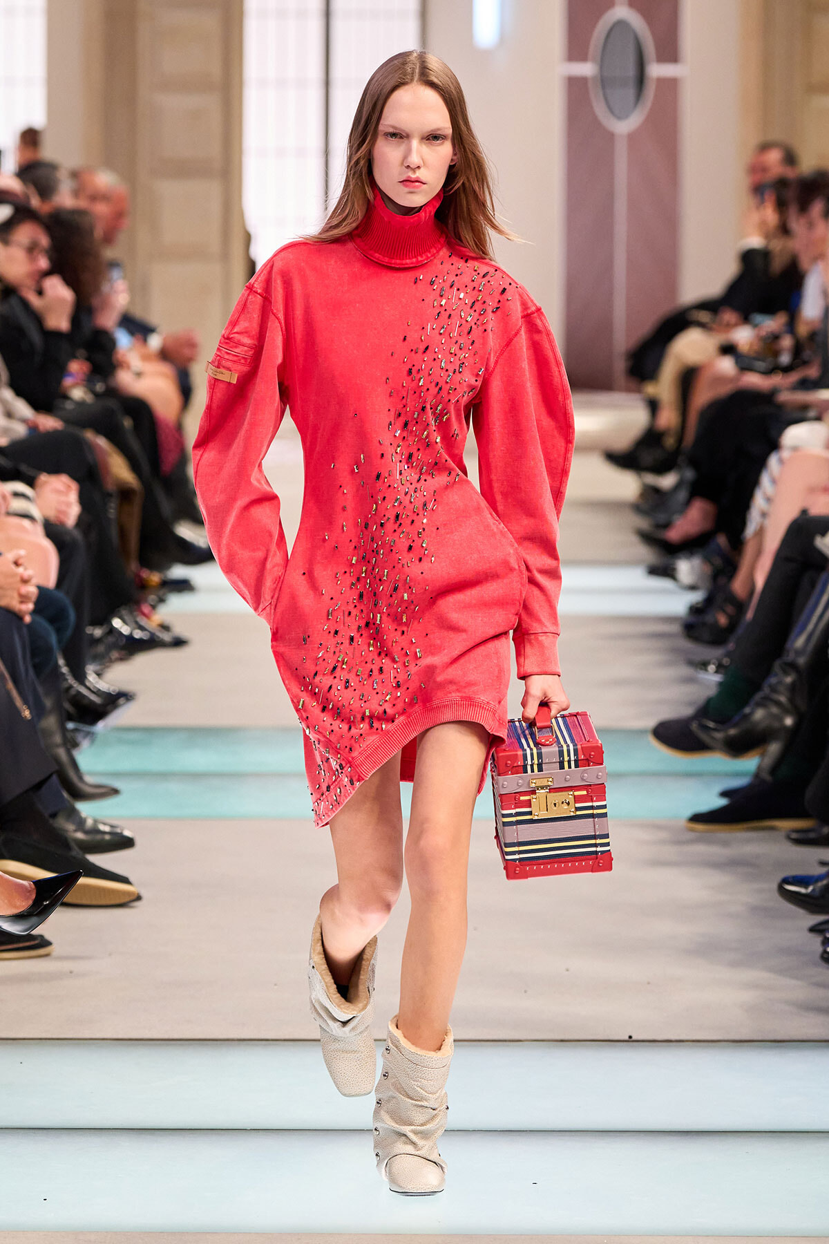 Model walking a fashion runway in a red turtleneck sweater dress with scattered black embellishments, carrying a striped box handbag and wearing light beige boots.