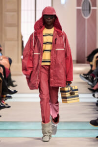 Runway model in a pink oversized parka with fur-lined hood, striped yellow sweater, pink pants, and silver slouch boots, carrying a yellow striped box bag.