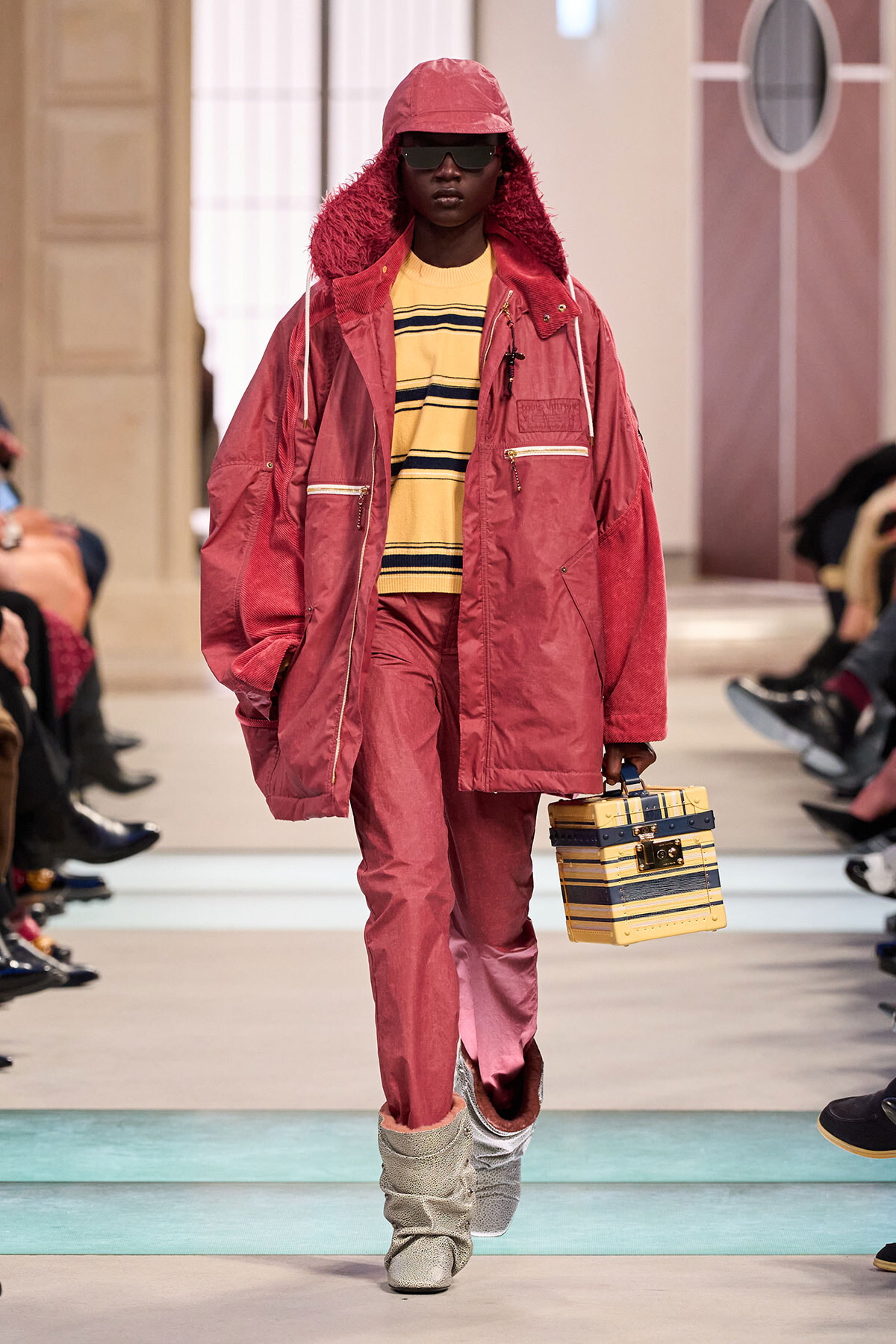 Runway model in a pink oversized parka with fur-lined hood, striped yellow sweater, pink pants, and silver slouch boots, carrying a yellow striped box bag.