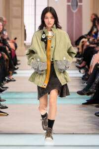 Model walking a runway in a oversized olive-green jacket over a mustard-orange top with a black-and-gray textured hem, onlookers in the background