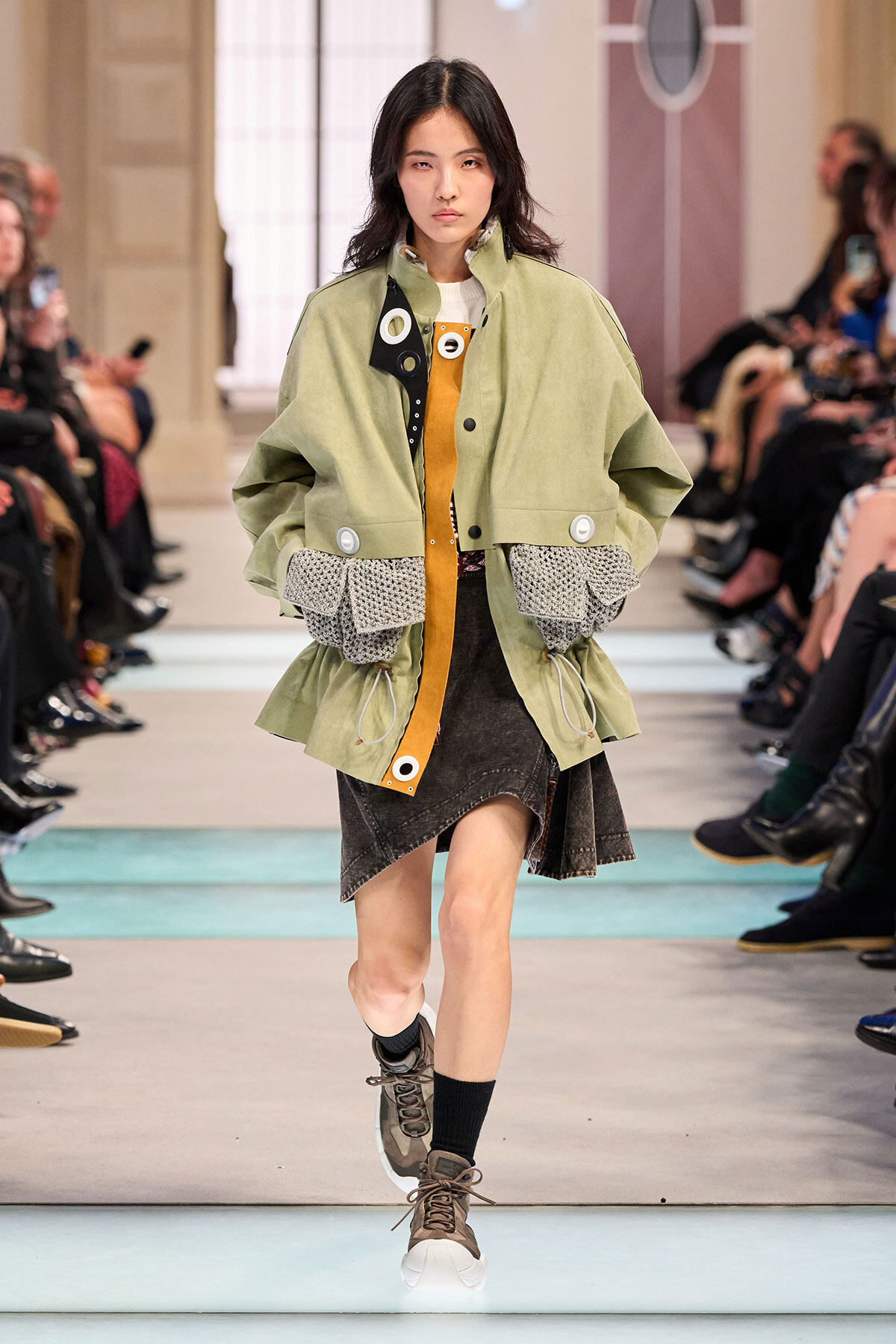 Model walking a runway in a oversized olive-green jacket over a mustard-orange top with a black-and-gray textured hem, onlookers in the background