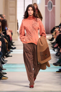 Model walking a runway in a peach turtleneck sweater with scattered embellishments and loose brown pants, holding a camel coat with a checkered pocket detail.