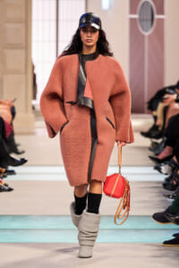 Model walking a runway in a oversized pink boucle coat with black leather accents, black cap, and a coral round bag.