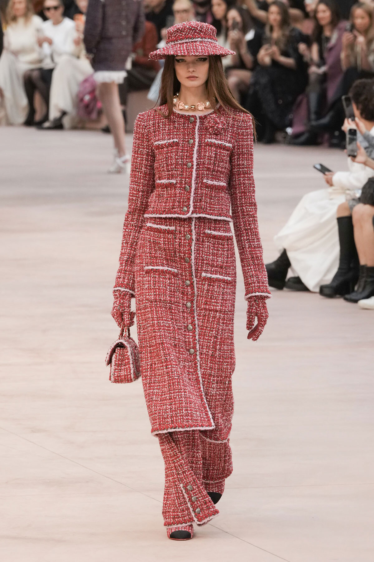 Model on a fashion runway wearing a red tweed suit with white trim, matching hat and gloves, carrying a small red handbag.