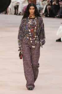 Model walks runway in a loose, multicolored plaid top with matching printed wide-leg pants and layered pearl necklaces, carrying a small burgundy handbag.
